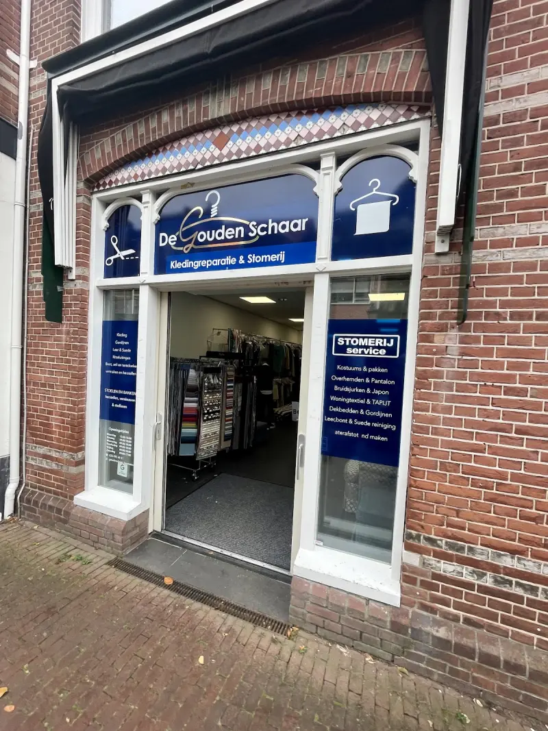 De winkelpui van De Gouden Schaar: witte kozijnen met blauwe ramen in een bakstenen gevel, de deur staat open